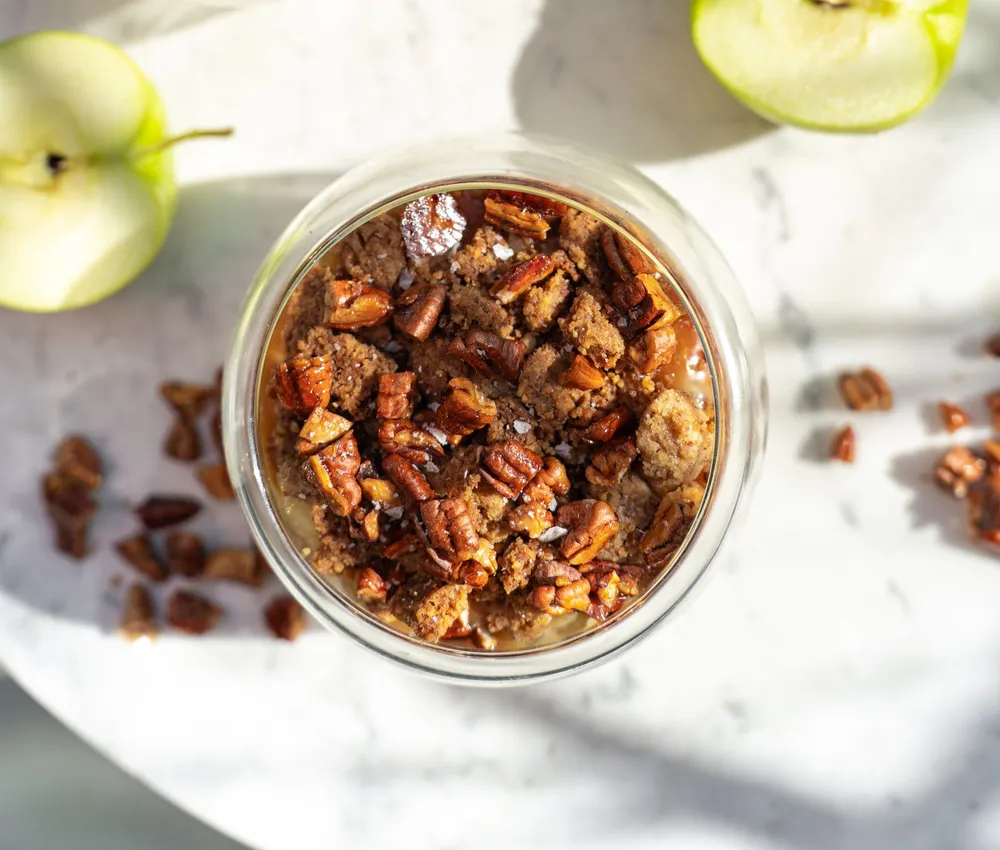Individual apple crumbles topped with browned crumbs and pecans, with green apples on a marble table