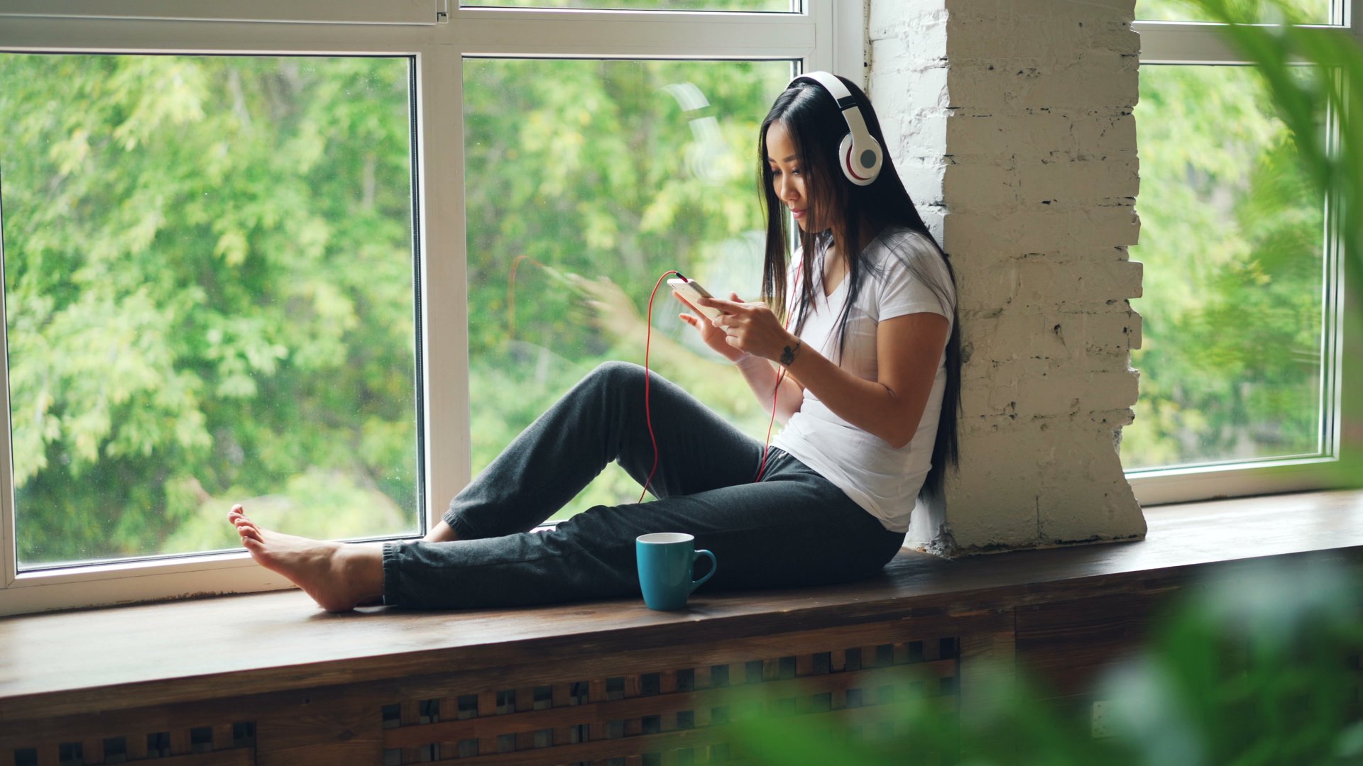 A woman sitting by a window with headphones and a phone